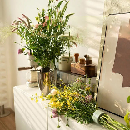 Vildblommor står i en blank metallvas på ett vitt skåp, medan en bukett med gula och rosa blommor ligger bredvid. 3