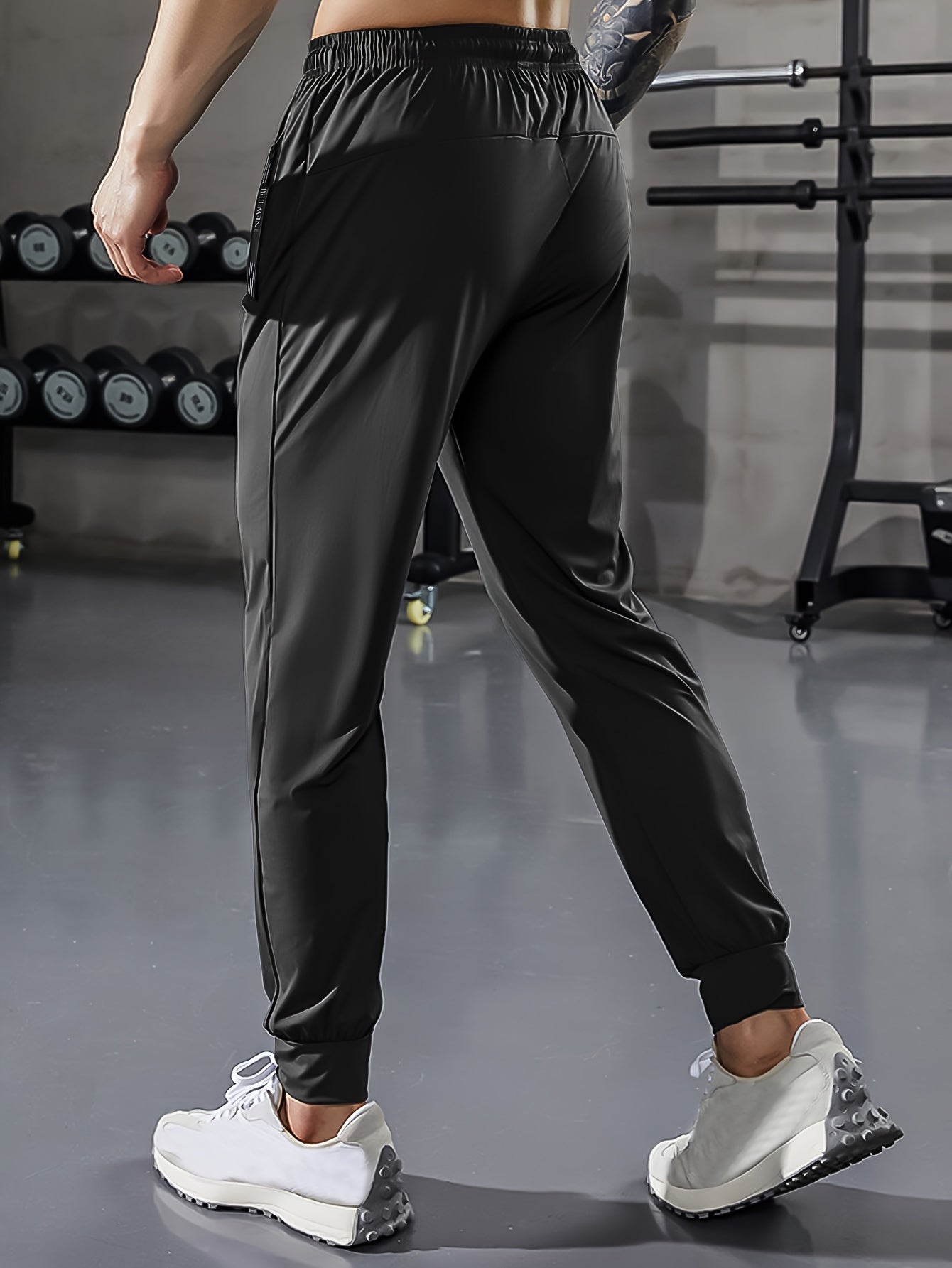 Mann in schwarzer Sporthose im Fitnessstudio, Hanteln im Hintergrund, weiße Turnschuhe.