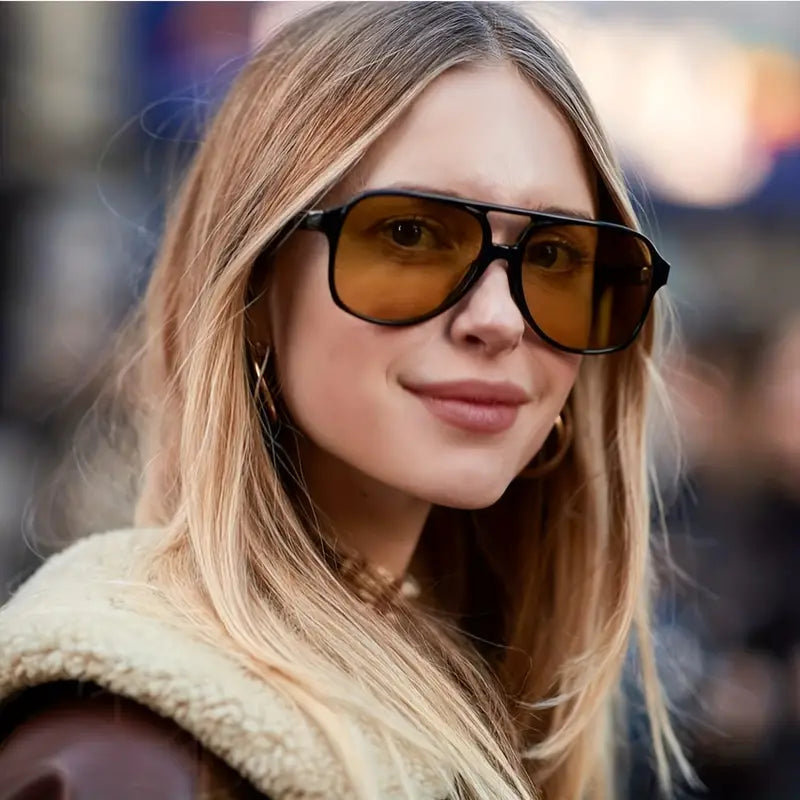 Frau mit blonden Haaren trägt große, eckige Sonnenbrille mit gelben Gläsern, modisch, urban.