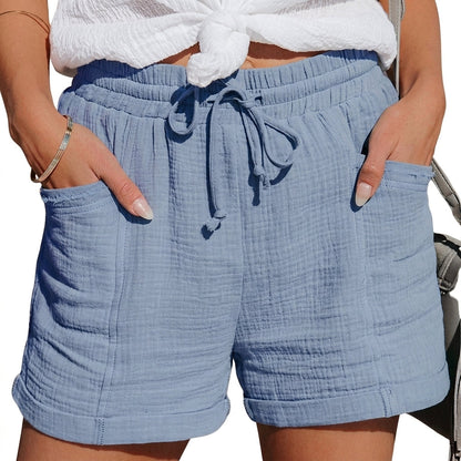 Modell i dusblå skrynklade shorts med knytsnöre, händerna i fickorna, vit knutad topp och grå väska. 1