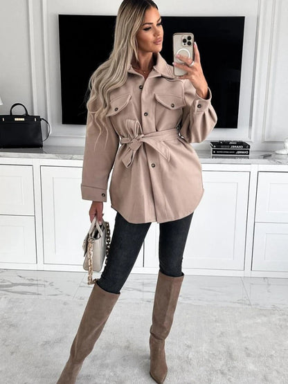 Frau in beiger Jacke mit Gürtel, schwarze Jeans, braune Stiefel, Smartphone, moderne Inneneinrichtung.