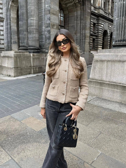 Frau in beiger Jacke und schwarzer Hose mit schwarzer Handtasche vor historischer Architektur.