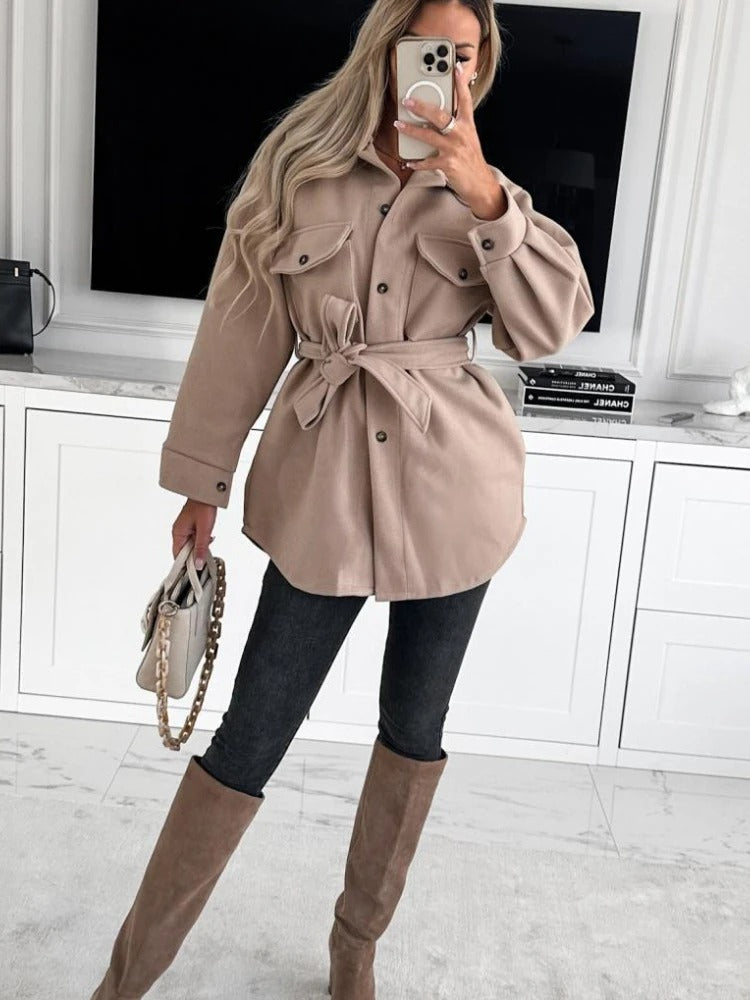 Frau in beiger Jacke mit Gürtel, schwarze Jeans, beige Stiefel, Handy, modisches Outfit.