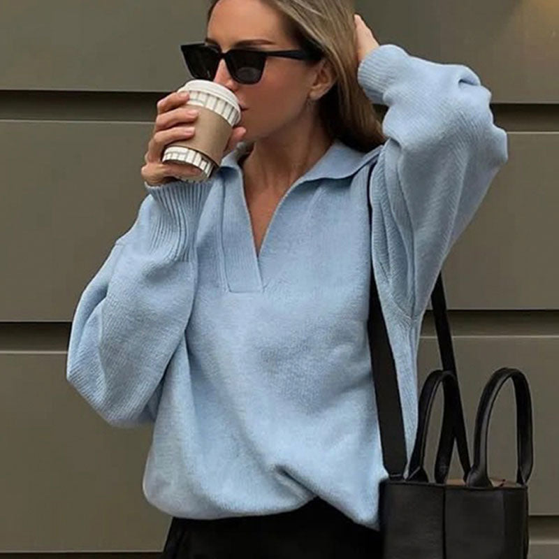 Frau in hellblauem Pullover trinkt Kaffee, trägt schwarze Sonnenbrille und Tasche.