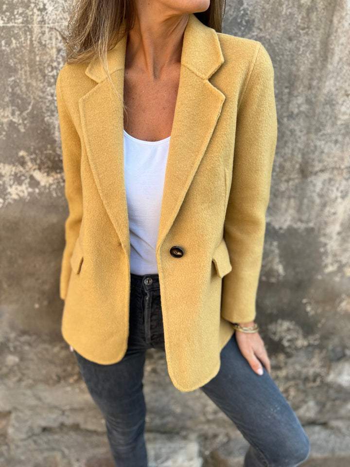 Frau in gelbem Wollblazer, weißem Top und Jeans vor einer Betonwand.
