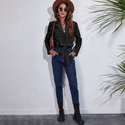 Frau in schwarzem Blazer, blauer Jeans, Hut und Stiefeln vor weißer Wand mit Pflanzen.