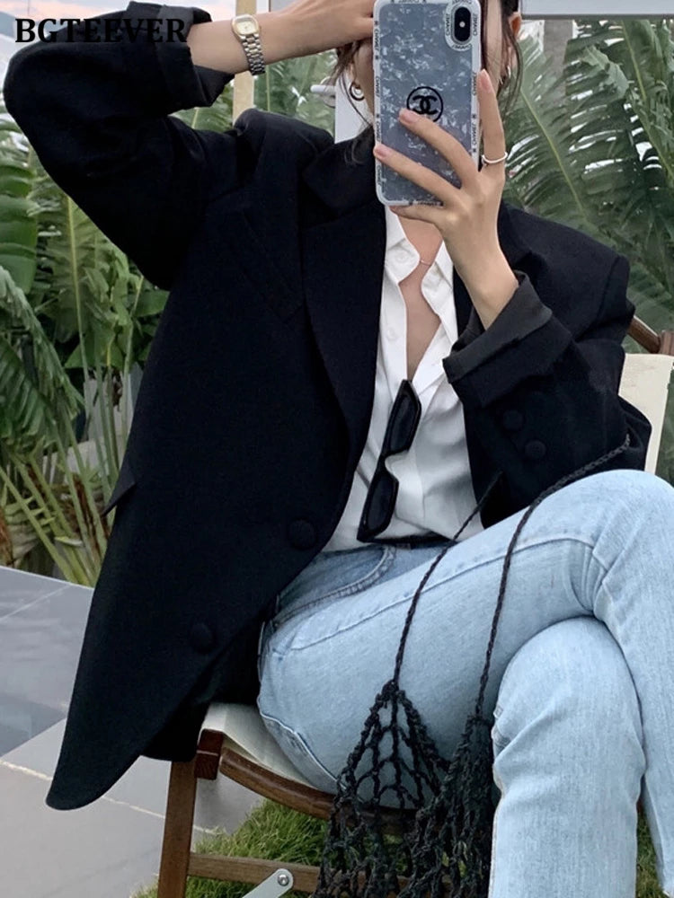 Frau in schwarzem Blazer, weißer Bluse, Jeans, hält Smartphone, sitzt draußen.