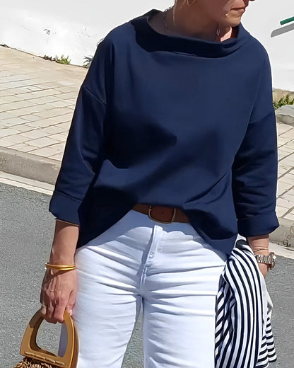 Frau in dunkelblauem Pullover und weißer Hose, hält gestreifte Jacke und Holzhandtasche.