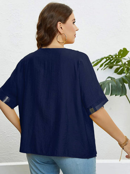 Frau in dunkelblauer Bluse mit kurzen Ärmeln, goldene Ohrringe, vor weißer Wand und Pflanze.