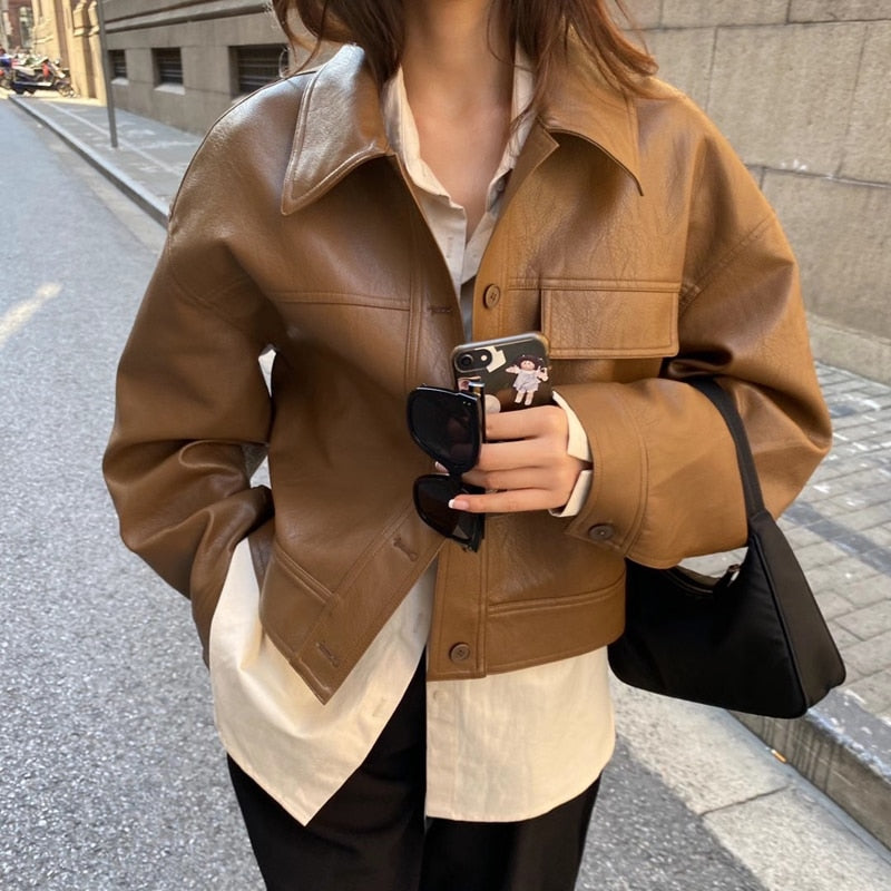 Frau in brauner Lederjacke, weißer Bluse, schwarze Hose, Smartphone, Sonnenbrille, Handtasche.