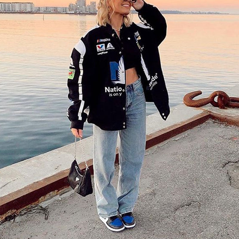 Frau in schwarzer Collegejacke, blauer Jeans und blauen Sneakers am Hafen bei Sonnenuntergang.