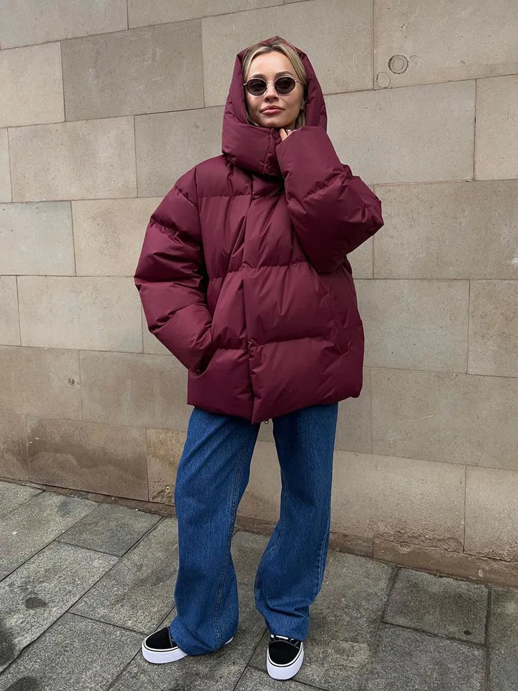 Frau in burgunderroter Daunenjacke, Jeans und Sonnenbrille vor Steinwand.