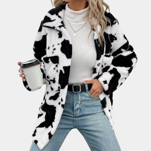 Frau in schwarz-weißer Kuhfelljacke, weißem Rollkragenpullover, Jeans, hält Kaffeebecher.