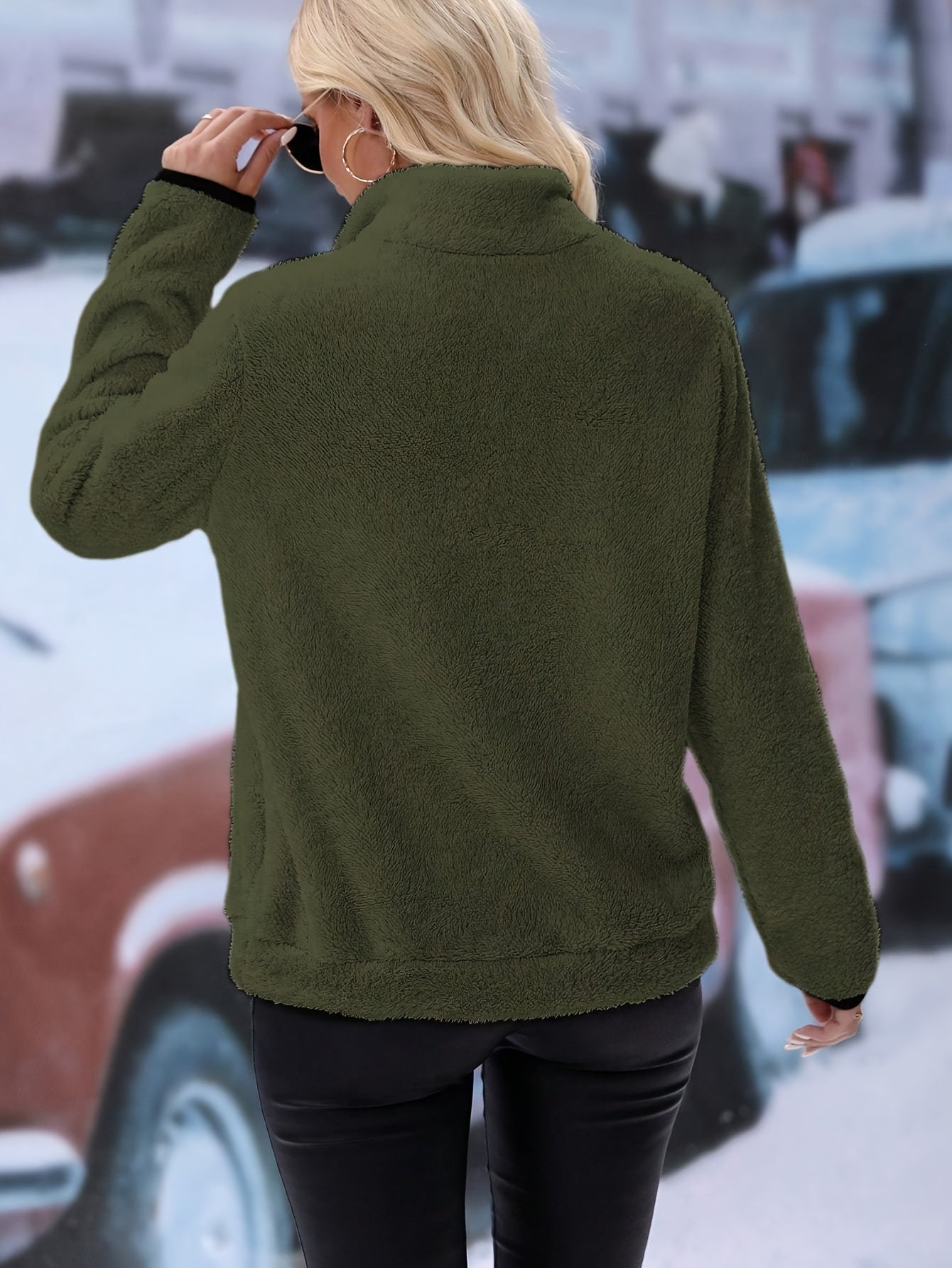 Frau in olivgrüner Fleecejacke, schwarze Lederhose, Sonnenbrille, Stadt im Hintergrund.