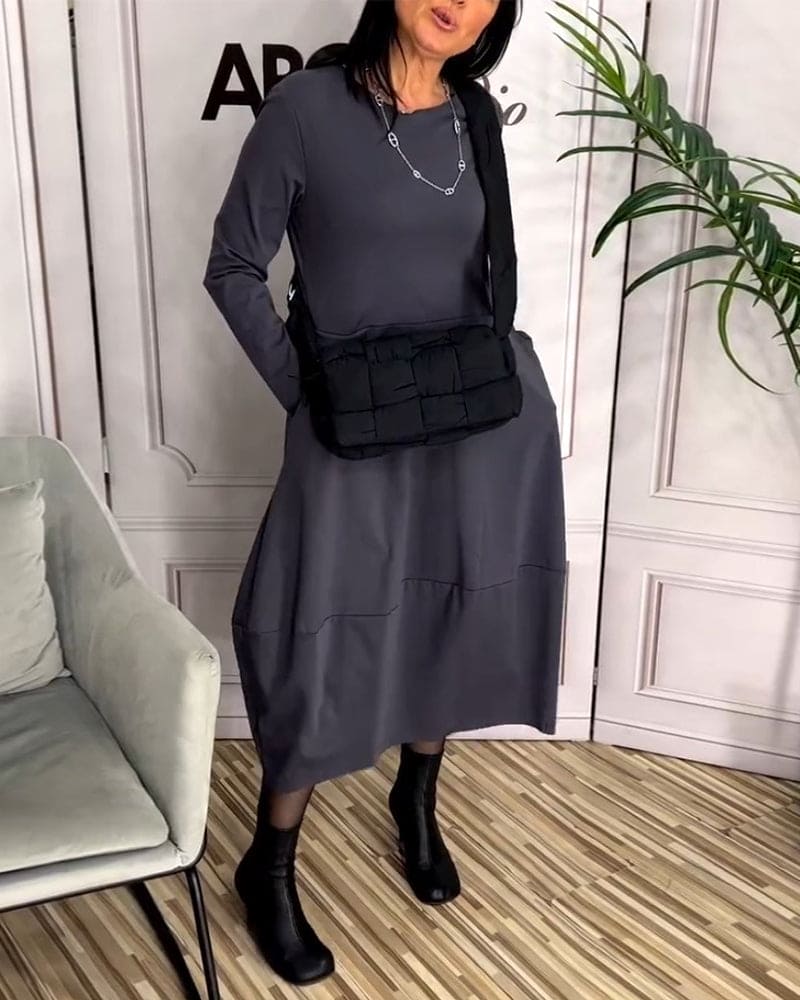 Frau in grauem Kleid mit schwarzer Umhängetasche und Stiefeln, steht in modernem Raum.