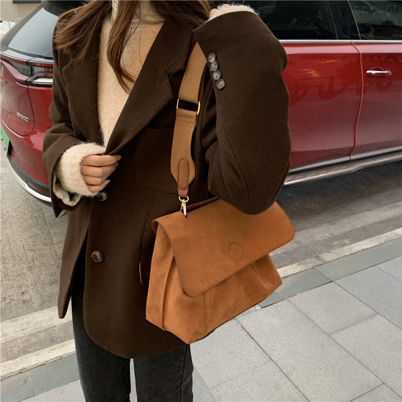 Frau mit brauner Wildledertasche, eleganter Mantel, rotes Auto im Hintergrund.