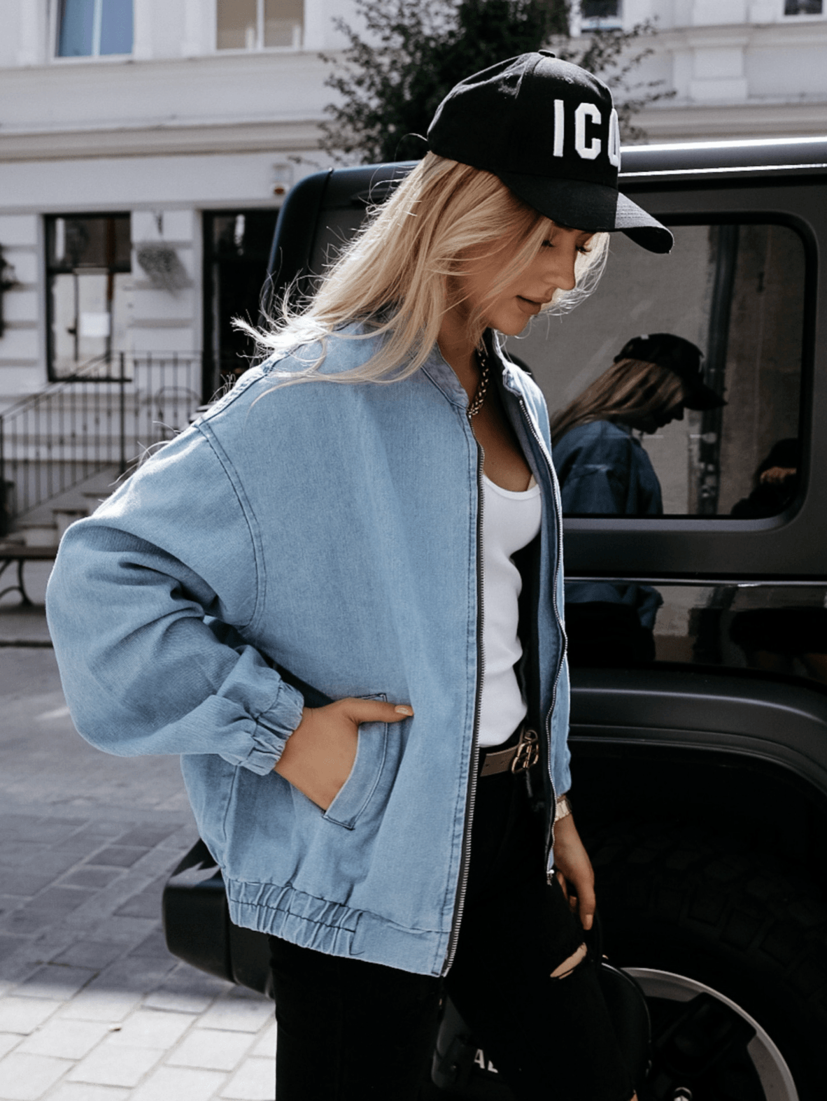 Frau in blauer Jeansjacke und schwarzer Kappe vor einem Auto auf der Straße.
