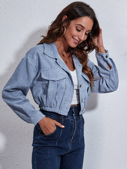 Frau in blauer Cordjacke und Jeans, lächelnd, vor weißer Wand.