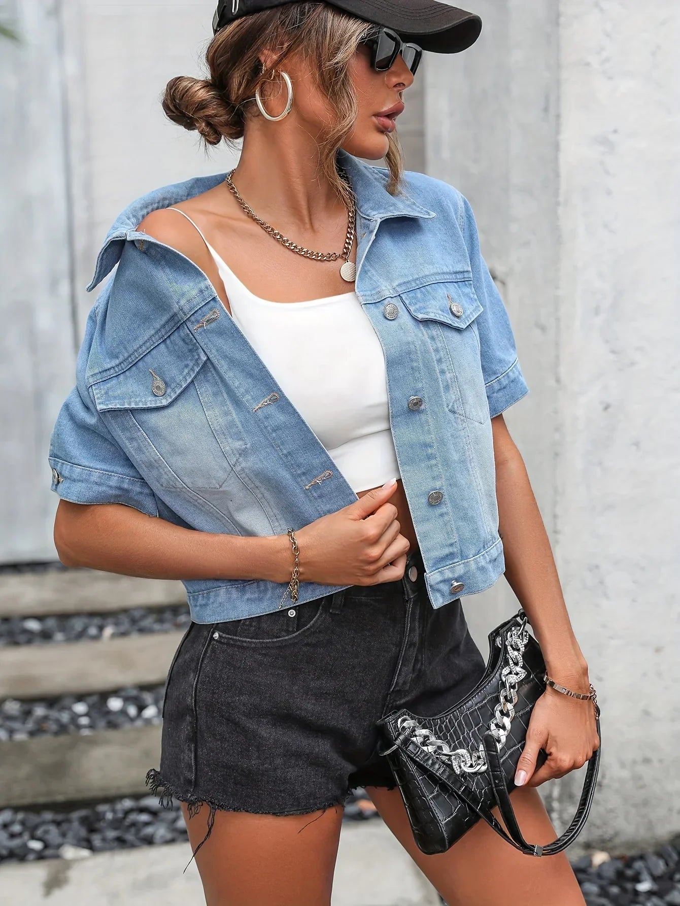 Frau in blauer Jeansjacke, weißem Top, schwarzer Shorts, Sonnenbrille und schwarzer Tasche.