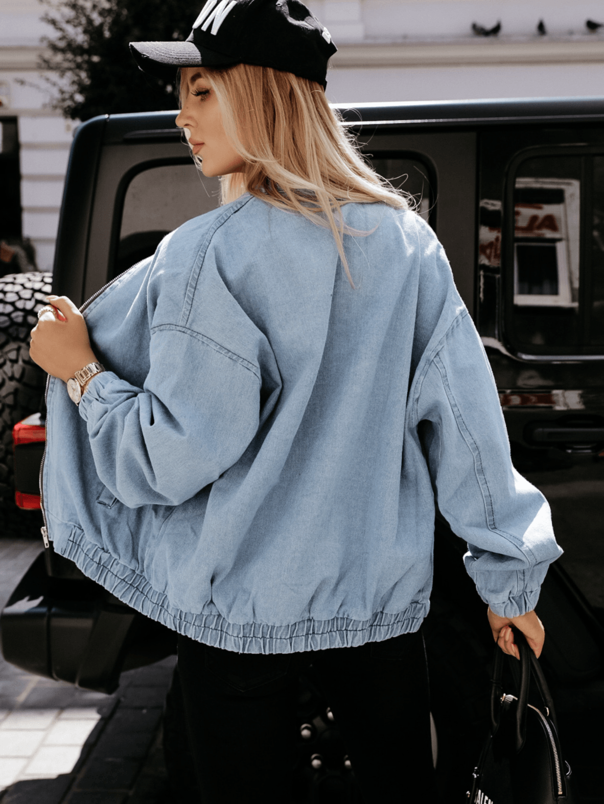 Frau in blauer Jeansjacke und schwarzer Kappe vor schwarzem SUV, modisch, urbaner Stil.