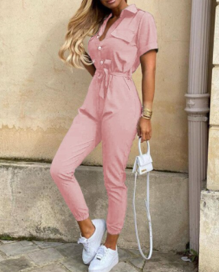 Frau in rosa Jumpsuit mit weißen Sneakers und kleiner weißer Handtasche vor beiger Wand.