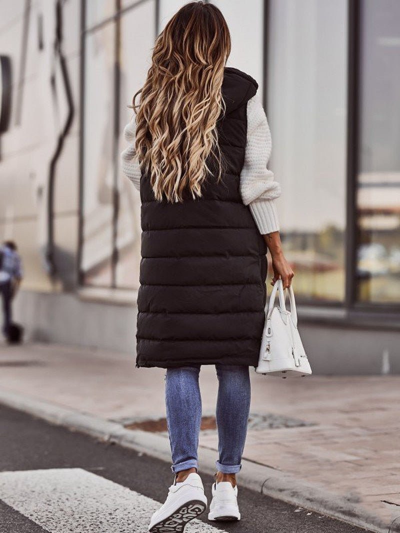 Frau in schwarzer, knielanger Steppweste, Jeans und weißen Sneakers auf Gehweg.