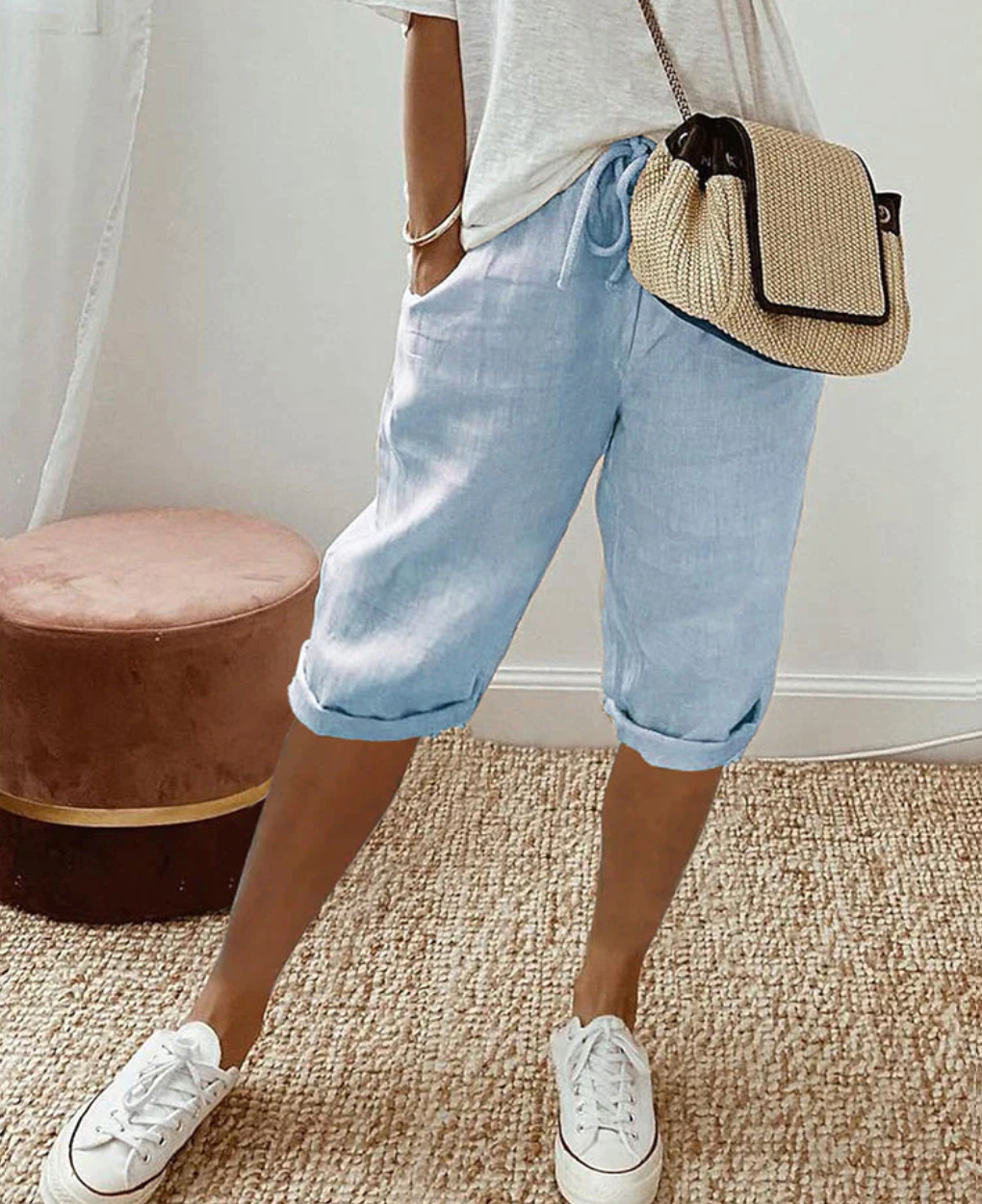 Frau in hellblauer Leinenhose, weißem T-Shirt, Strohtasche, weiße Sneaker, Sommermode.