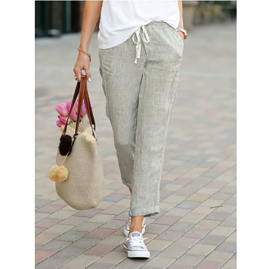 Frau in Leinenhose, weißem T-Shirt, Sneakers, trägt Strohtasche mit Pompons, Straße.