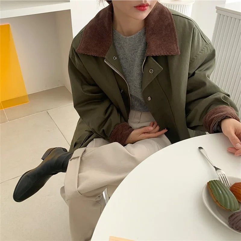 Frau in olivgrüner Jacke mit braunem Kragen, grauem Pullover, beiger Hose, sitzt am Tisch.