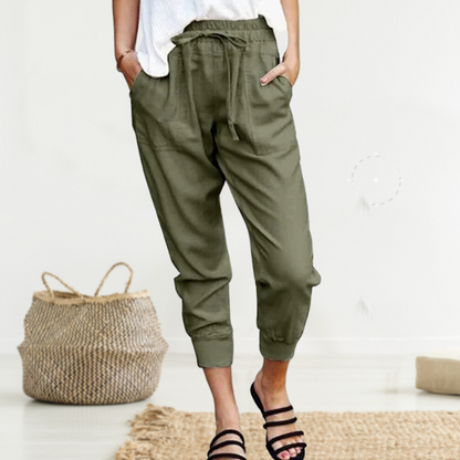Frau in olivgrüner Leinenhose, weißem Oberteil, schwarze Sandalen, moderner Wohnraum.