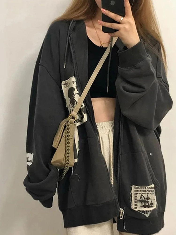 Frau in übergroßer, grauer Kapuzenjacke mit Aufnähern, beige Umhängetasche, lässiger Stil.