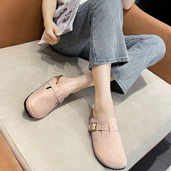 Frau trägt rosa Wildleder-Clogs mit Schnalle, sitzt auf Sofa, lässige Jeans.