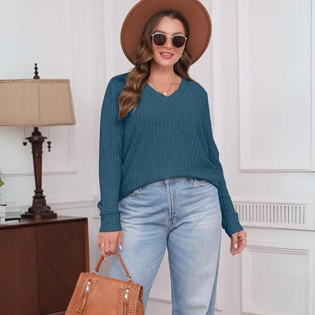 Frau in blauem Pullover, Jeans, braunem Hut und Sonnenbrille, steht in modernem Wohnzimmer.