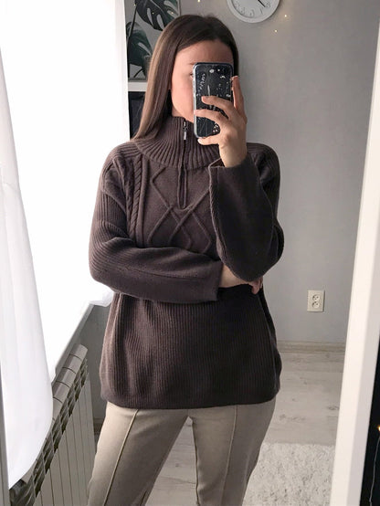 Frau in braunem Strickpullover mit Reißverschluss, beige Hose, modernes Interieur.