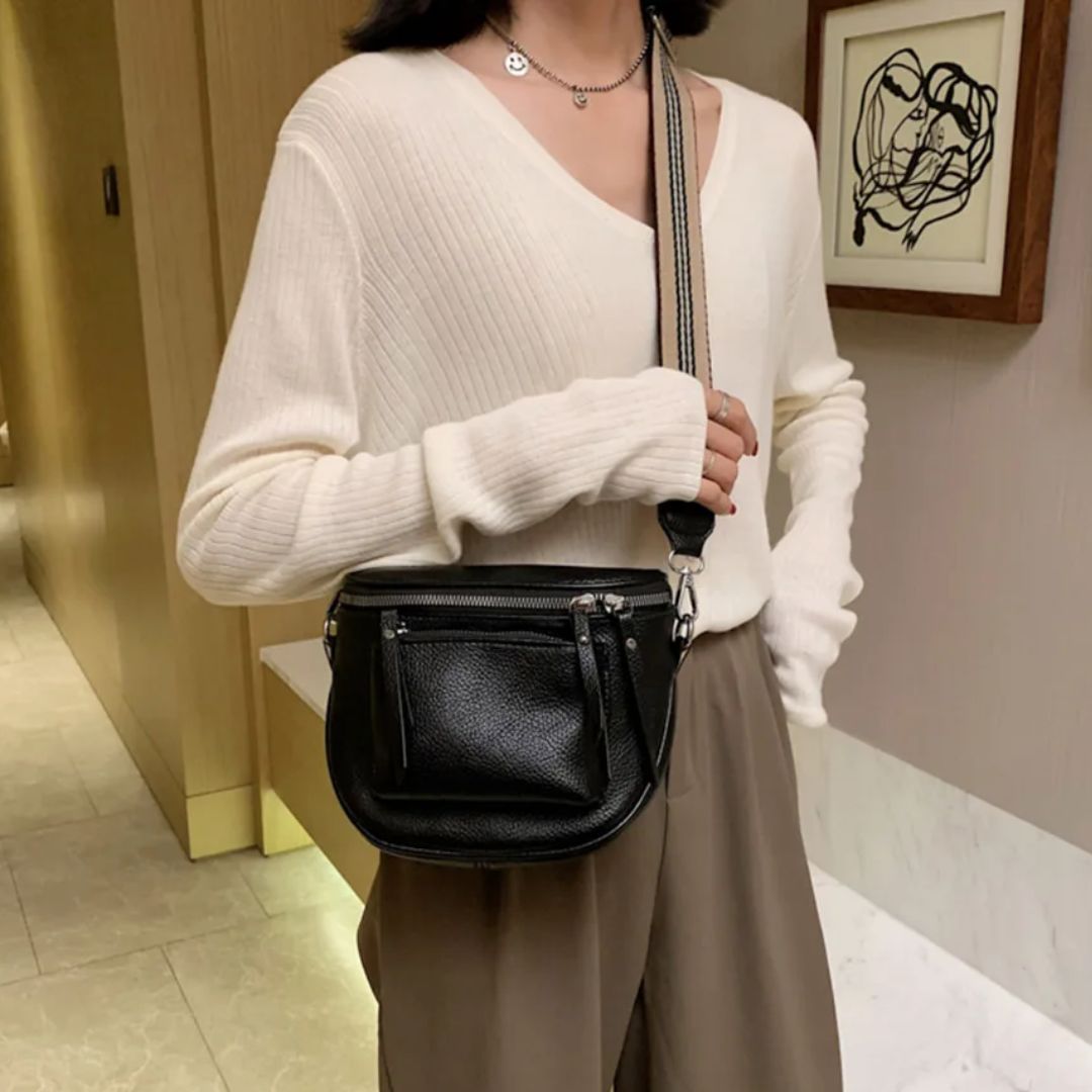 Frau mit schwarzer Leder-Umhängetasche, cremefarbenem Pullover, brauner Hose in modernem Raum.