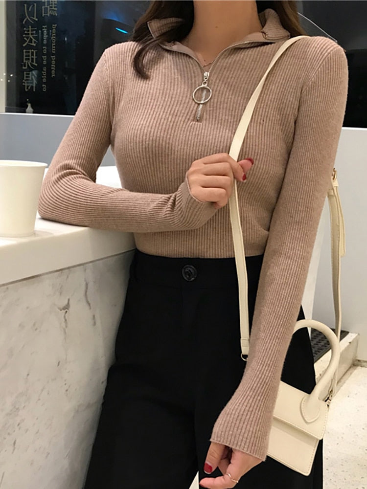Frau in beige geripptem Pullover mit Reißverschluss, schwarze Hose, weiße Umhängetasche.