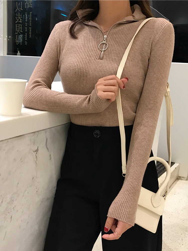 Frau in beige geripptem Pullover mit Reißverschluss, schwarze Hose, weiße Umhängetasche.