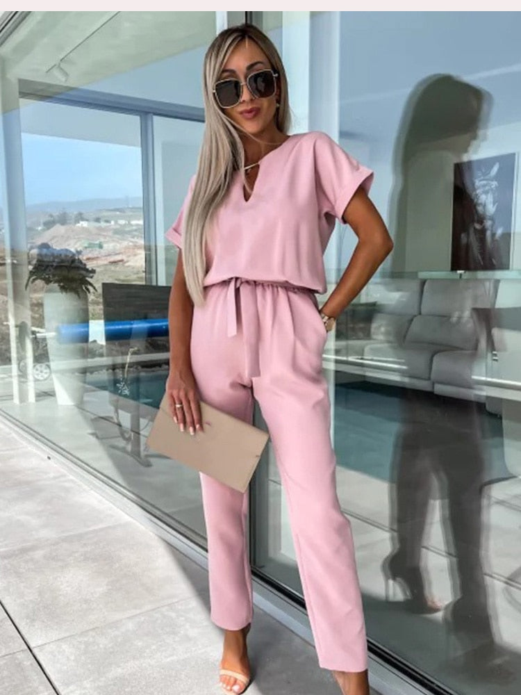 Frau in rosa Jumpsuit mit Sonnenbrille, beige Clutch, moderne Terrasse im Hintergrund.