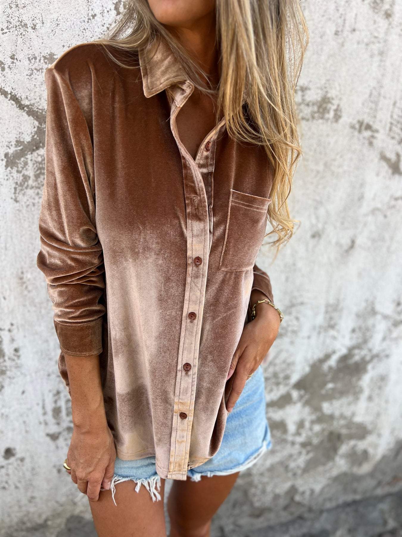 Frau in brauner Samt-Hemdbluse und Jeansshorts vor Betonwand.