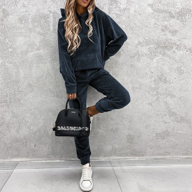 Frau in dunkelgrauem Samt-Hoodie und Jogginghose, hält schwarze Balenciaga-Tasche, weiße Sneaker.