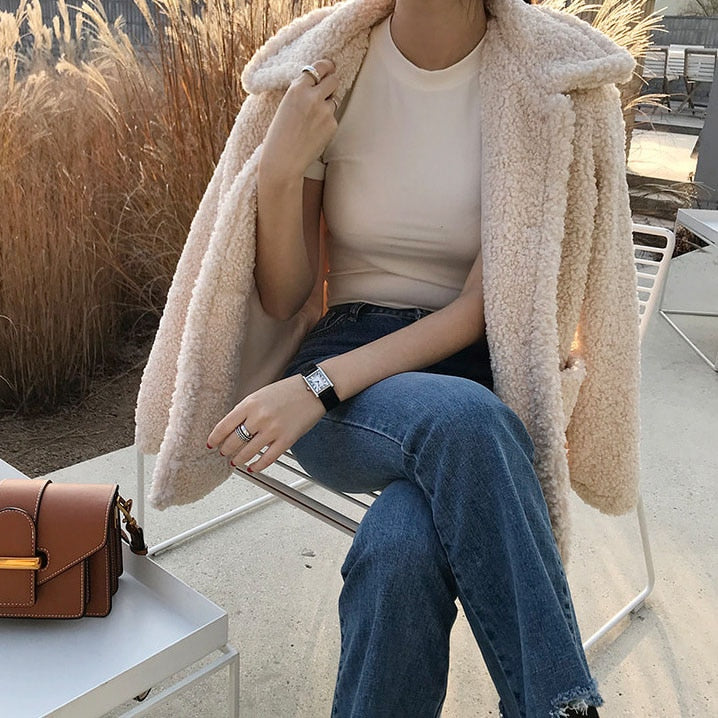 Frau in weißem T-Shirt und beiger Teddyjacke, sitzt auf Stuhl, braune Tasche, Jeans.