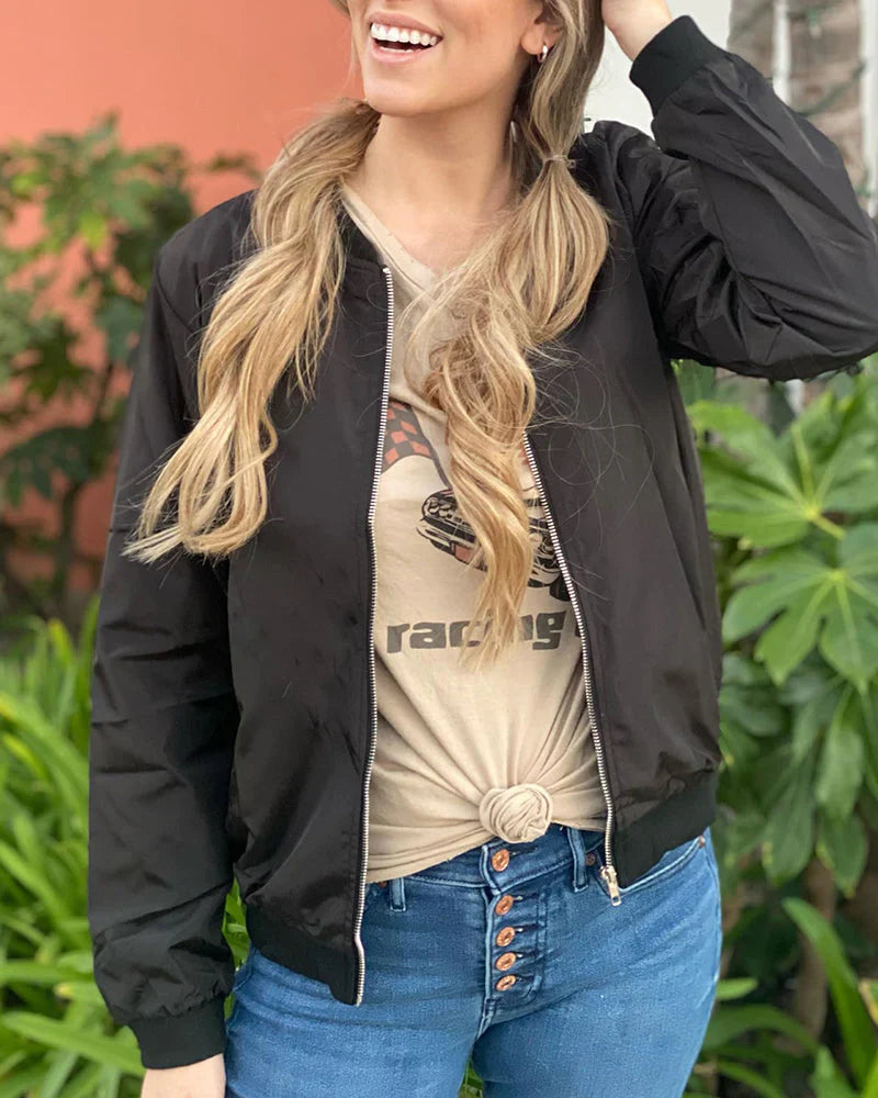 Frau in schwarzer Bomberjacke, beige T-Shirt, blaue Jeans, lächelnd im Freien.