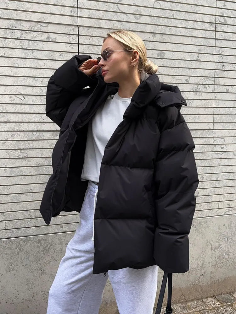 Frau in schwarzer Daunenjacke, weiße Jogginghose, Sonnenbrille, vor grauer Wand.