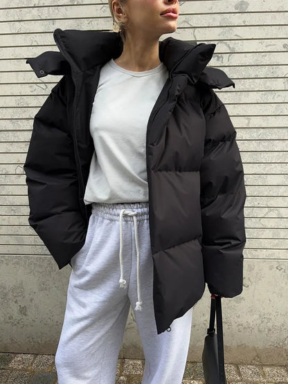 Frau in schwarzer Daunenjacke und weißem Sweatshirt vor Betonwand.