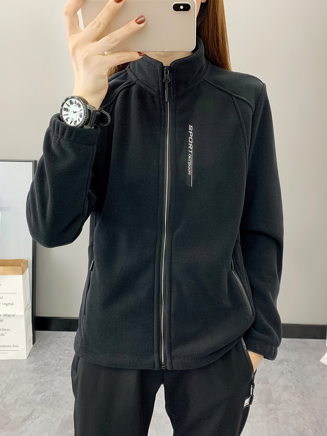 Frau in schwarzer Fleecejacke mit Reißverschluss, Sportuhr, Smartphone, Indoor-Hintergrund.