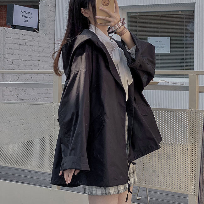 Frau in schwarzer Kapuzenjacke, kariertem Rock, weißer Bluse, urbaner Hintergrund.