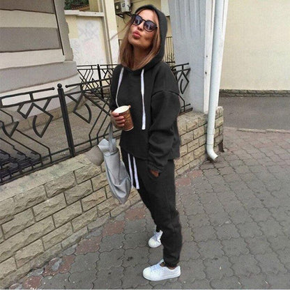 Frau in schwarzem Hoodie und Jogginghose, Sonnenbrille, hält Kaffeebecher, lässiger Streetstyle.
