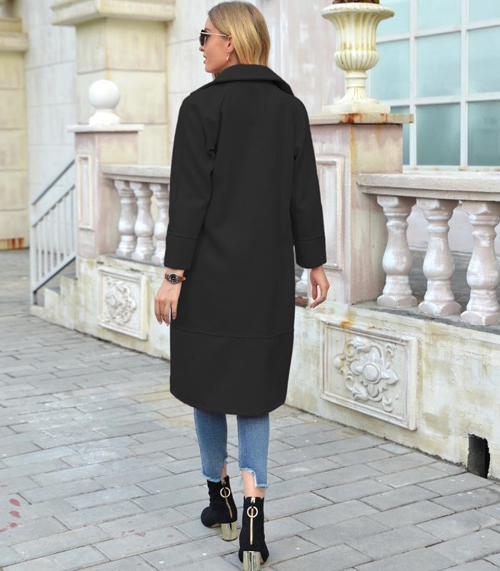 Frau in schwarzem Mantel, Jeans und Stiefeletten auf gepflastertem Gehweg vor Gebäude.