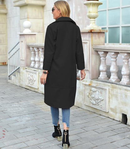 Frau in schwarzem Mantel, Jeans und Stiefeletten auf gepflastertem Gehweg vor Gebäude.