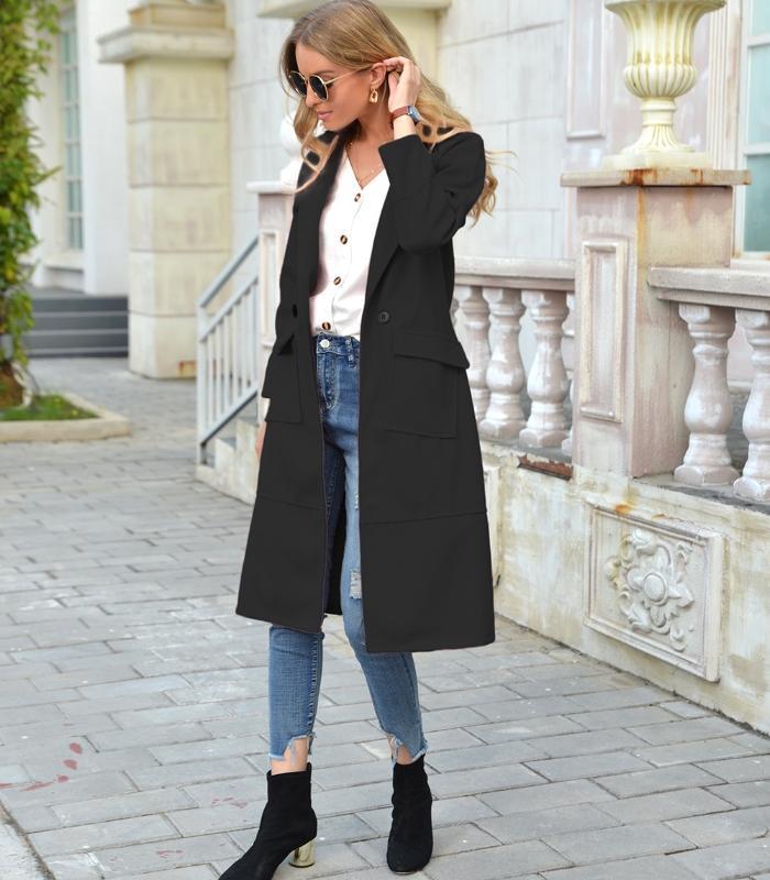 Frau in schwarzem Mantel, Jeans und Stiefeletten auf Gehweg vor Gebäude.